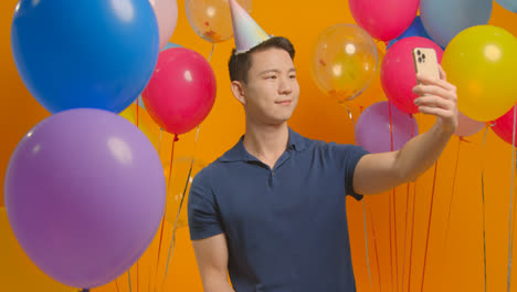 Studioporträt-Eines-Mannes,-Der-Ein-Selfie-Mit-Partyhut-Macht-Und-Seinen-Geburtstag-Feiert,-Umgeben-Von-Luftballons